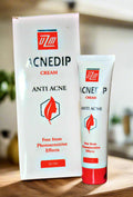 Two tubes of Acnedip anti-acne cream on a wooden surface with a blurred background.